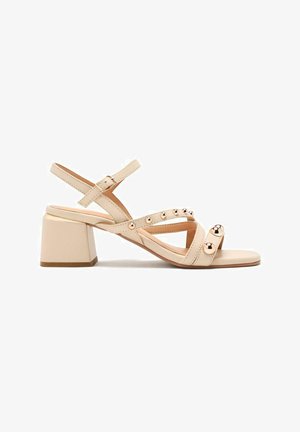 Sandales à talons en beige avec un talon carré, dotées de brides croisées et d'accents métalliques ronds. Texture lisse et bride de cheville ajustable.