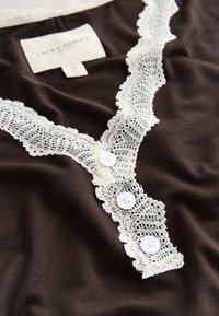 Dunkelbraunes Stoffoberteil mit weißer Spitzenborte entlang des Ausschnitts und drei weißen Knöpfen, zeigt ein Laura Ashley Bekleidungslabel.