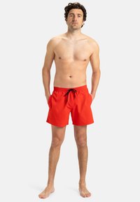 Costume de baie roșu dintr-un material ușor, dotat cu un brâu elastic și un șnur negru, precum și cu buzunare laterale.