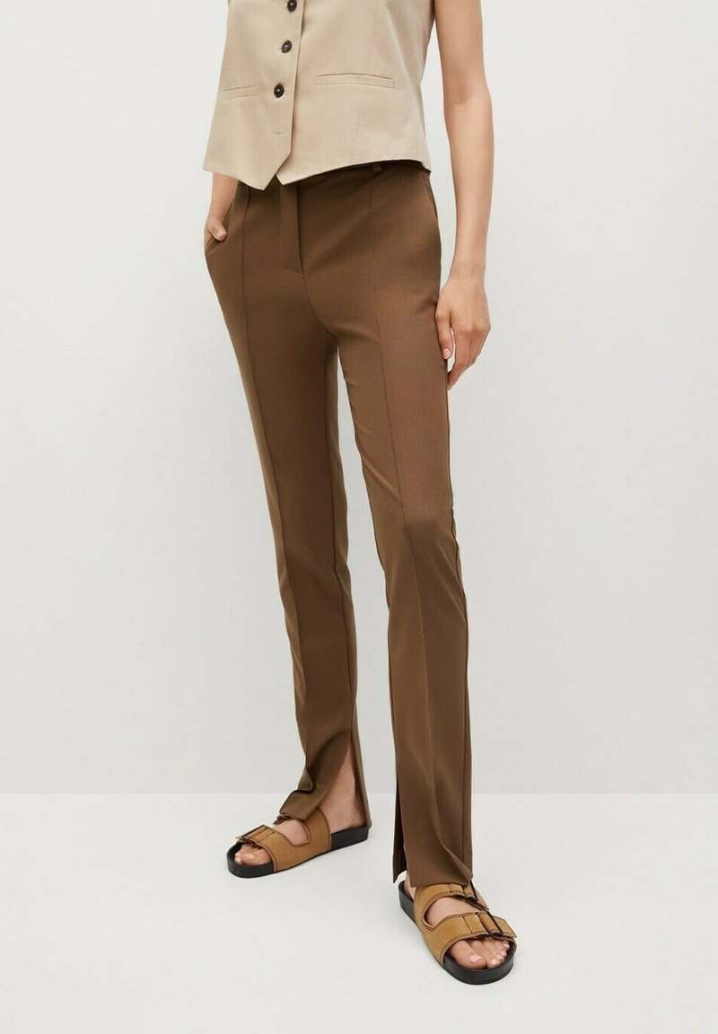 Personne portant un pantalon marron slim avec des fentes à l'avant des ourlets, un gilet beige sans manches avec des boutons, et des sandales marron à bouts ouverts.