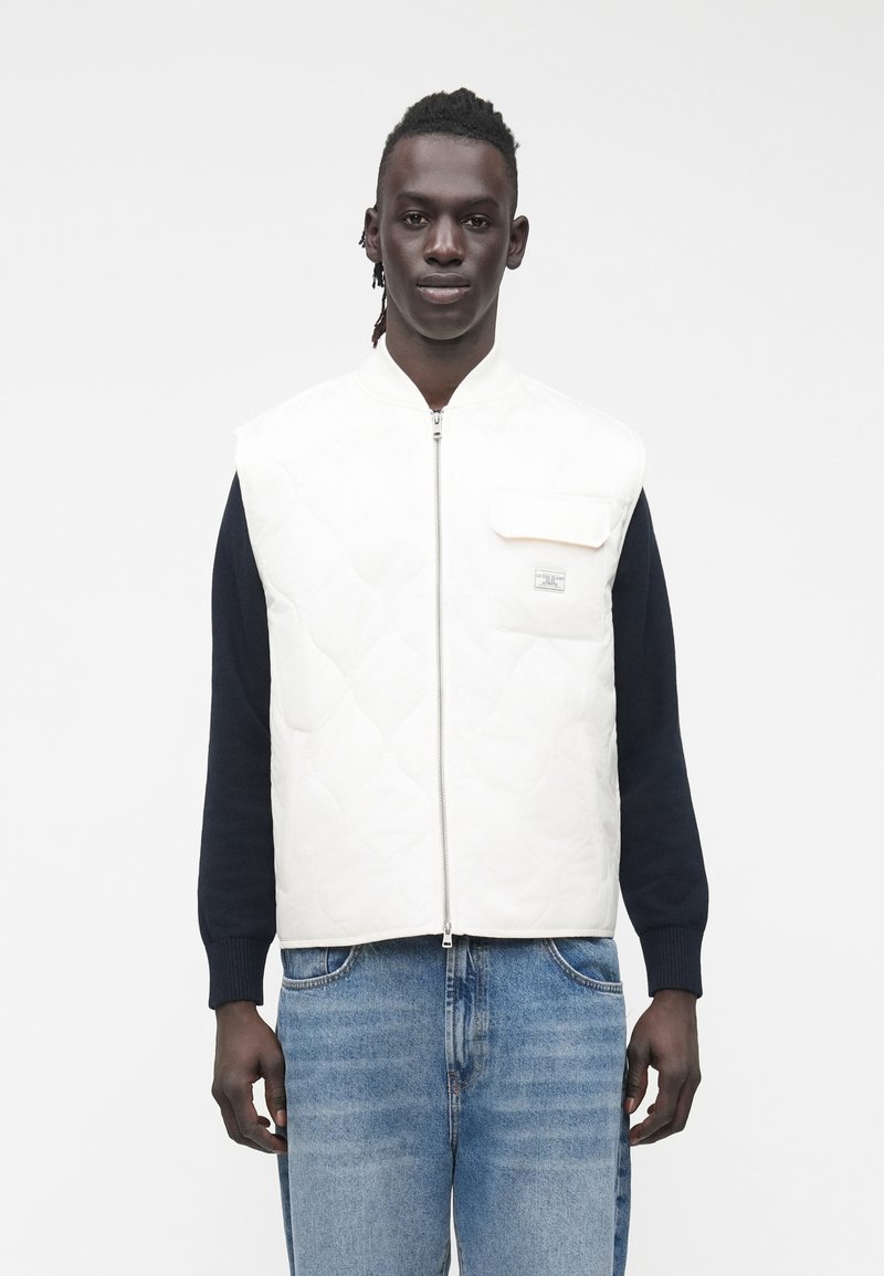 Juodas vyras dėvi baltą siuvinėtą liemenę su krūtinės kišenėle per tamsiai mėlyną ilgomis rankovėmis marškinius ir šviesiai mėlynas džinsus, stovintis priešais baltą foną.