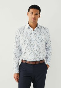 Bărbat îmbrăcat într-o cămașă albă cu mânecă lungă și un model floral albastru, pantaloni închiși la culoare și o curea maro, stând cu o mână în buzunar.
