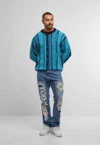 Maglione turchese a trecce con motivi blu e neri, abbinato a jeans chiari strappati con vari buchi e toppe colorate.