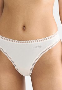 String en coton blanc avec une bordure en dentelle à motif festonné, texture lisse et ceinture subtilement marquée.