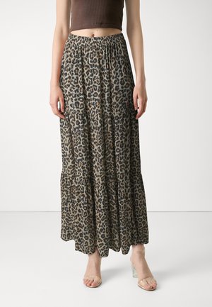 Vrouw draagt een maxi-rok met luipaardprint en een elastische taille, gecombineerd met beige hakken met transparante bandjes, staand tegen een effen witte achtergrond.