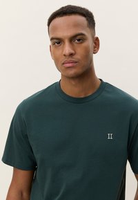 Σκούρο πράσινο βαμβακερό T-shirt με στρογγυλή λαιμόκοψη και κοντά μανίκια, που διαθέτει κεντημένο λογότυπο "II" σε λευκό χρώμα στο αριστερό στήθος.