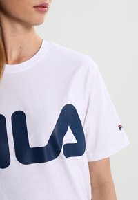 Bílý bavlněný tričko s velkým námořnickým modrým logem "FILA", krátkými rukávy a malým akcentem loga na pravém rukávu.