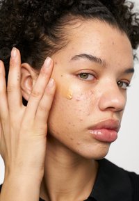Modèle appliquant un fond de teint beige sur sa joue. Sa peau présente une texture et un aspect imparfait. Elle a les cheveux bouclés et une expression neutre.