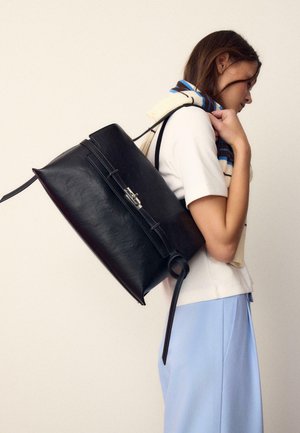 Ženska nosi veliko črno usnjeno torbo čez ramo, oblečena v belo kratkoročno majico, modro krilo in črtasto rutko, ki ji je prek ramen.