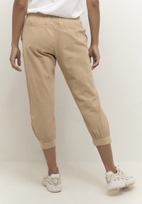 Bež kratke hlače iz lahkotnega materiala z elastičnim pasom in zavihanimi robovi. Oblečene s kremnimi supergami.