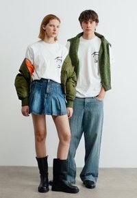 Δύο νεαροί ενήλικες στέκονται δίπλα δίπλα, φορώντας λευκά μπλουζάκια με σχέδια. Ο ένας φοράει πράσινο μπουφάν και τζιν φούστα με μαύρες μπότες, ενώ ο άλλος φαρδιά τζιν παντελόνια και πράσινο γιλέκο.