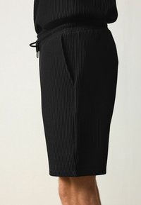 Pantalones cortos de canalé negros con cinturilla elástica y cordón, que cuentan con bolsillos laterales y un diseño de longitud hasta la rodilla. Detalle de tejido texturizado visible.