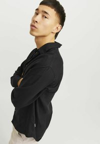 Camicia nera a maniche lunghe in una vestibilità comoda con colletto a punta. Realizzata in tessuto leggero, presenta una texture sottile e un dettaglio con etichetta laterale.