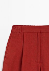 Pantaloni rossi in lino con vita elastica, dotati di pieghe frontali e tasche laterali. Il tessuto ha una finitura textured.