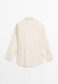 Blouse beige à manches longues avec col, couture en yoke au dos, poignets boutonnés et détail de cordon de serrage à la taille, présenté sur un fond blanc.