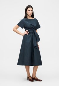 Γυναίκα στέκεται με το χέρι στη μέση φορώντας ένα ναυτικό μεσάτο φόρεμα με κοντά μανίκια, δεμένη μέση και καφέ παπούτσια Mary Jane.