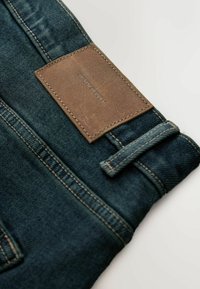Tumši zili džinsa džinsi ar teksturētu brūnu ādas plāksteri ar uzrakstu "DENIM SUPPLY." Ir kontrastējošas šuves un jostas cilpa.