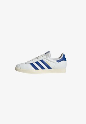 White leather sneaker with blue stripes, round toe, flat lace-up design, textured rubber sole, and yellow "GAZELLE" branding.