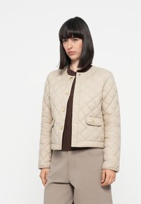 Quilted beige jacket with a round neckline, gold buttons, and side pockets, paired with a brown sweater and light gray pants.