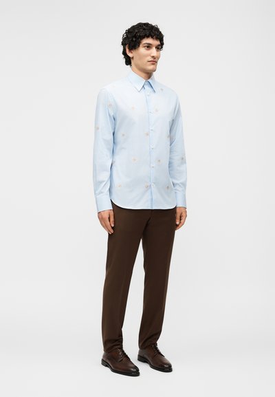 Άνδρας που στέκεται, φοράει ανοιχτό μπλε πουκάμι με μακριά μανίκια με μικρά ανθισμένα μοτίβα, καφέ παντελόνι και σκούρα καφέ επίσημα παπούτσια πάνω σε λευκό φόντο.