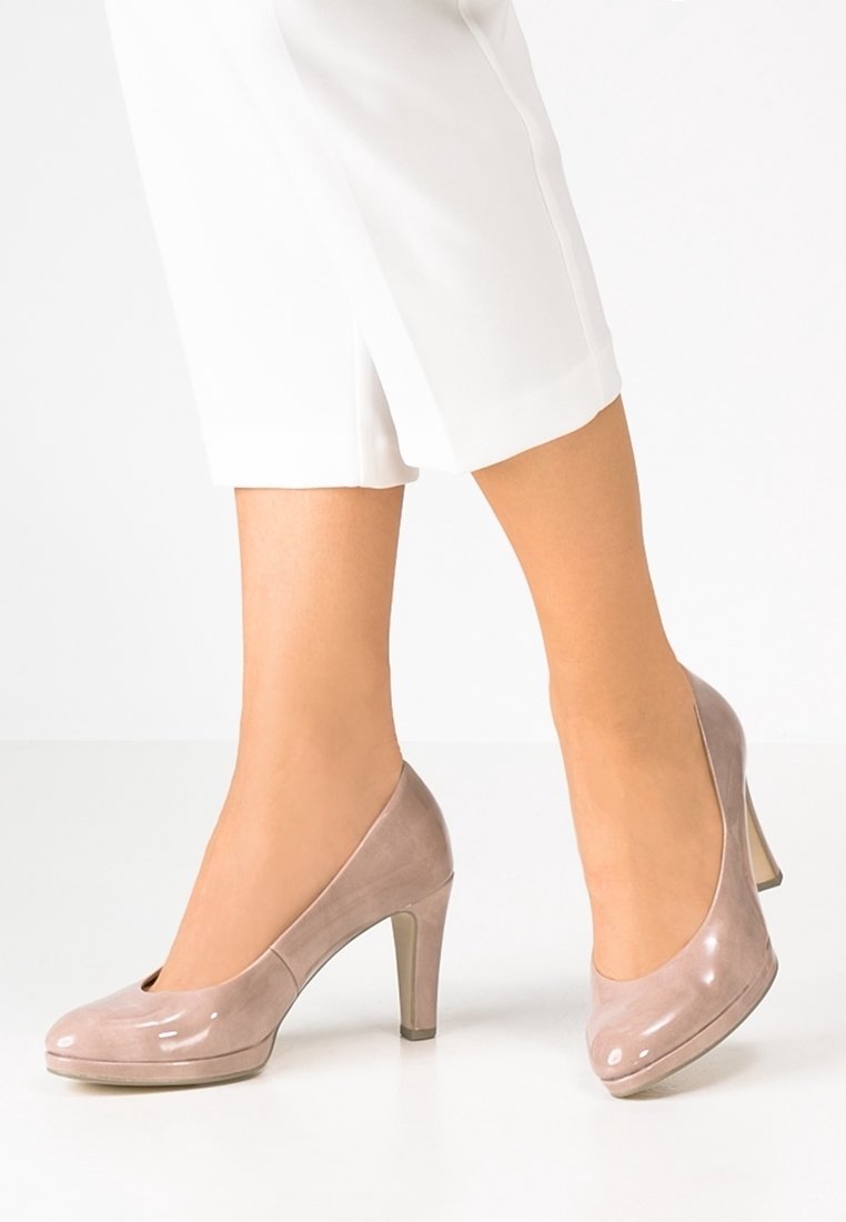 Jambes portant un pantalon blanc raccourci et des chaussures beige à talons hauts en cuir verni, debout sur un sol blanc.