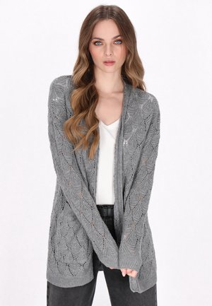 Grijze gebreide cardigan met een bladerenpatroon, open voorkant, lange mouwen en ribgebreide manchetten. Draagt over een witte top en donkere jeans.