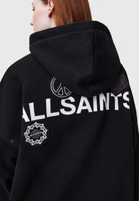 Sudadera negra con capucha, que presenta el texto blanco "ALLSAINTS" y varios parches bordados, incluyendo un símbolo de paz y un diseño de cadena.