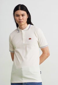 Krémové polo tričko s rebrovaným golierom, ktorý má fialové pruhy. Malá logo nášivka na hrudi. Krátke rukávy a mäkká bavlnená textúra.