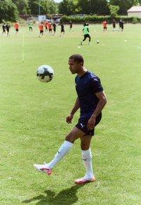 Atlet v tmavě modrém dresu od Pumy s bílými akcenty, který balancuje černobílým fotbalovým míčem na noze, na zelené trávě s tréninkem v pozadí.