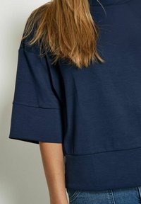 Top cropped bleu marine avec des manches larges et une texture lisse. L'ourlet a une coupe droite, associé à un jean en denim léger.