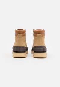 Bottes en daim beige avec des accents en cuir marron, semelles en caoutchouc noir côtelées et bout arrondi. Col court et rembourré pour plus de confort.