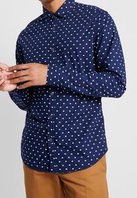 Chemise bleu marine à boutons, avec un motif rectangulaire blanc, manches longues et ourlet arrondi. Conçue en tissu lisse, elle arbore un col classique et des poignets.