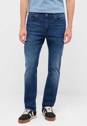 Mann trägt blaue Slim-Fit-Jeans, hellblaues T-Shirt und schwarze Sneaker mit weißen Schnürsenkeln und steht vor einem einfarbigen Hintergrund.