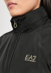 Zwarte zip-up jas van een glad, glanzend materiaal met een hoge kraag en gouden logo-details op de borst.