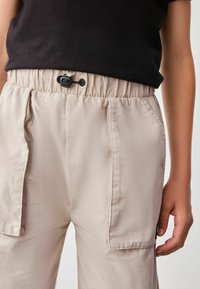Beige Hosen mit elastischem Bund und verstellbarem Kordelzug, ausgestattet mit Seitentaschen und einer glatten, leichten Textur.