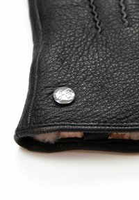 Schwarze Lederhandschuhe mit einem silbernen Druckknopf und geprägten Initialen. Weiches Futter sichtbar am Öffnungsbereich, mit feinen Nähdetails.