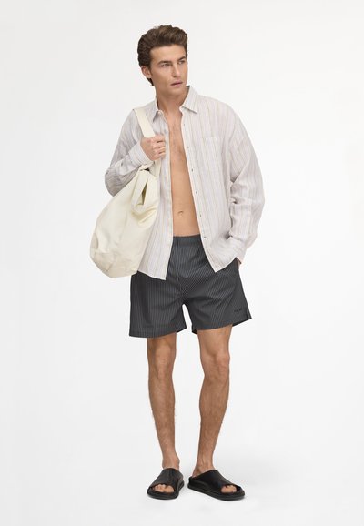 Jeune homme portant une chemise à rayures ouverte, un short noir à fines rayures, des sandales noires, portant un sac fourre-tout blanc, debout sur fond blanc.