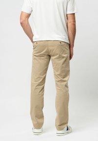 Vyras dėvi smėlio spalvos kelnes, baltus sportbačius ir baltą trumpomis rankovėmis palaidinę, stovintis su rankomis kišenėse, atsukti nugara.