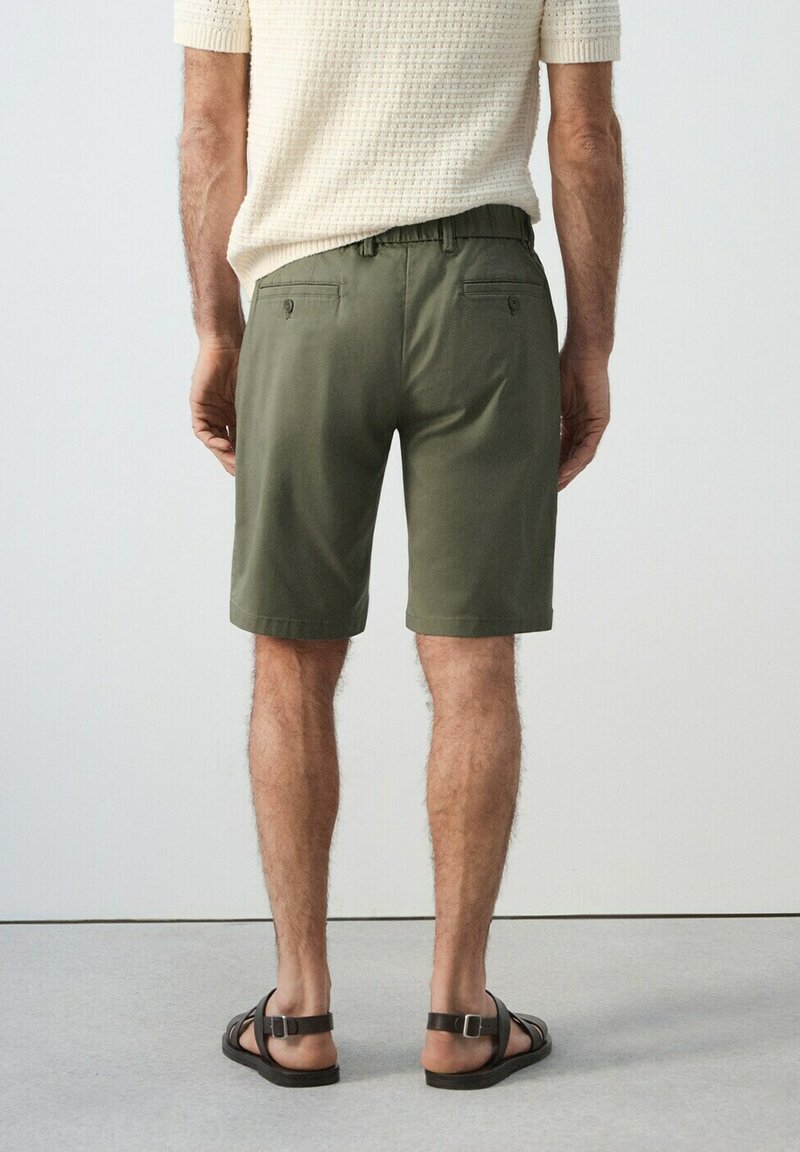 Homme portant un short vert olive, un haut en maille crème à manches courtes et des sandales noires, debout de dos sur un fond uni.