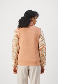 Fleeces jaka ar mīkstu persiku krāsas aizmuguri un rakstainām krēma krāsas piedurknēm, kurās ir ģeometriski raksti zemes toņos. Augsts apkakle un brīvas piegriezuma siluets.