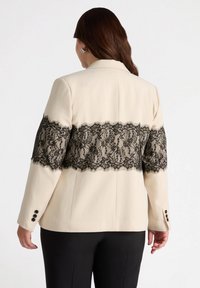 Blazer beige con un panel de encaje negro en la cintura, con patrones detallados. Incluye botones negros en las mangas.