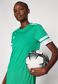 Groen sportshirt met witte accenten, korte mouwen en een ronde hals. Houdt een zwart-wit gestreepte voetbals bal vast.