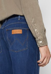 Jeans de mezclilla azul oscuro con un parche de cuero marrón de Wrangler y un patrón de costura distintivo en la parte trasera, combinados con una camisa de pana beige.
