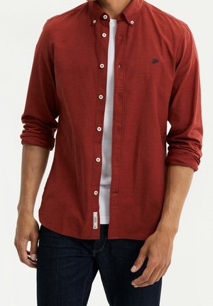 Homme portant une chemise rouge boutonnée par-dessus un t-shirt blanc avec les manches retroussées et un jean bleu foncé.