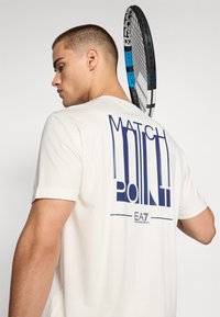 Balta trumpa rankovė t-shirt su mėlyna grafinė tekstu "MATCH POINT" ant nugaros, su lygiu paviršiumi. Matomas teniso raketė.