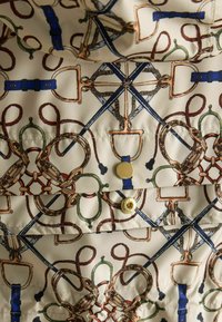 Kremveida audums ar drukātu jāšanas dizainu, kas ietver virves, bitus, jostus un zelta pogas uz quiltēta tekstila virsmas.