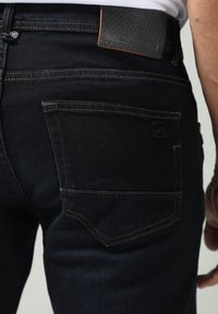 Jeans de mezclilla oscura con un parche de cinturilla de cuero negro texturizado, acentos en naranja y bolsillos traseros estándar con sutiles detalles de costura.