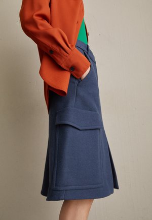 Osoba ubrana w niebieską spódnicę do kolan z dużą klapką kieszeni oraz pomarańczową, długą koszulę, z ręką włożoną do kieszeni, stojąca na tle jednolitej ściany.