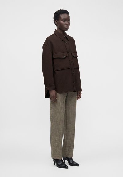 Femme portant une veste marron foncé boutonnée avec de grandes poches avant, un pantalon droit à carreaux et des bottines noires à talons, debout sur un fond blanc.