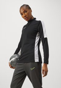 Zwarte sporttop met lange mouwen en witte zijpanelen, met een ritskraag, gecombineerd met grijze broek met een groen Nike-logo, terwijl er een zwart-witte bal vastgehouden wordt.
